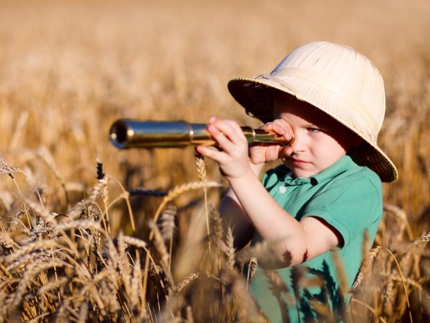 bambino con cappello da esploratore in mezzo ad un campo di spighe di grano guarda attraverso un binocolo