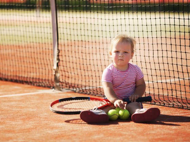 bambino piccolo seduto su un campo da tennis con in mano racchetta e palline