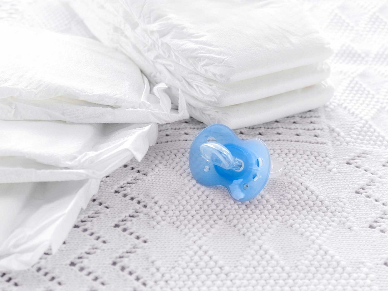 ciuccio blu riposto su lenzuolo ricamato bianco a fianco di pannolini nuovi