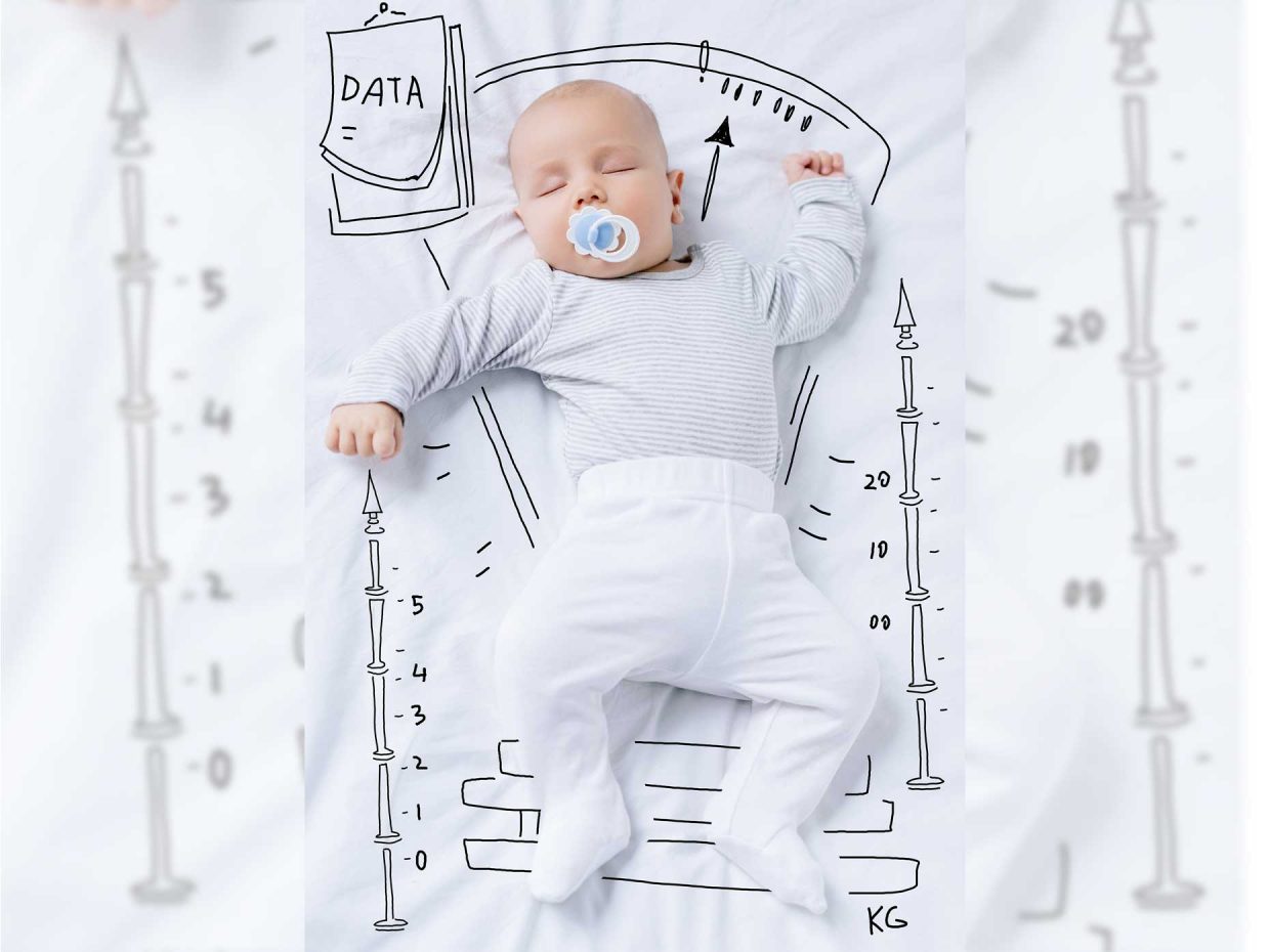 neonato con ciuccio in bocca è disteso su una coperta ornata da disegni che riproducono una bilancia