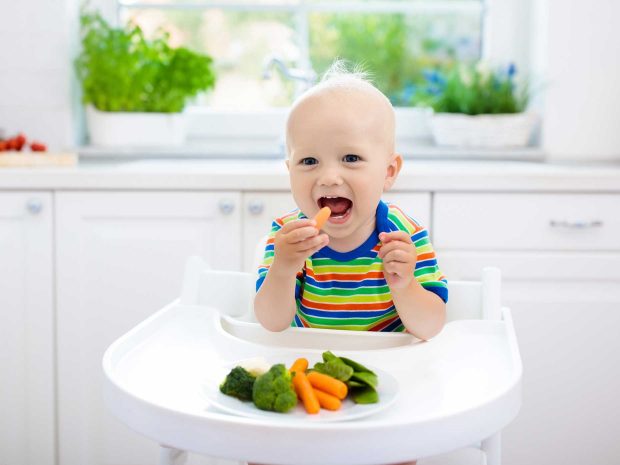 bambino seduto sul seggiolone mangia delle carote e dei broccoletti